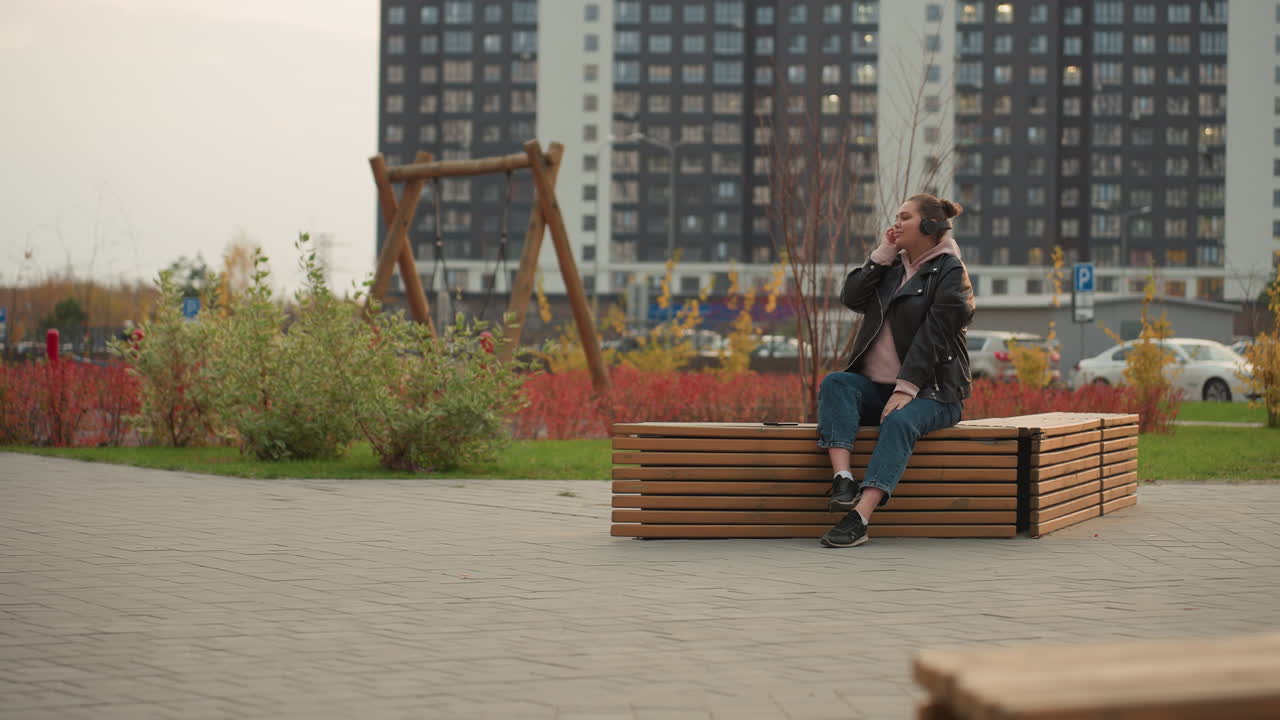 Woman seated on wooden bench in park listens to music with headphones while shrubs sway gently in wind surrounded by urban scene featuring parked cars tall buildings and street sign in background
