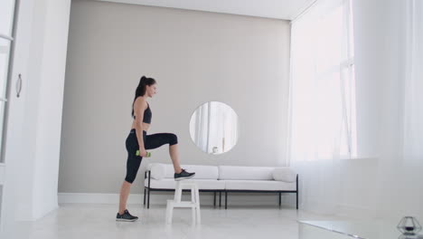 una joven morena realiza un paso concentrado en una silla en un apartamento de interior brillante con pesas en las manos.