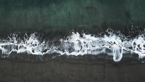 a wave hitting the black sand beach, showcasing the contrast of white foam against the dark shoreline, aerial view