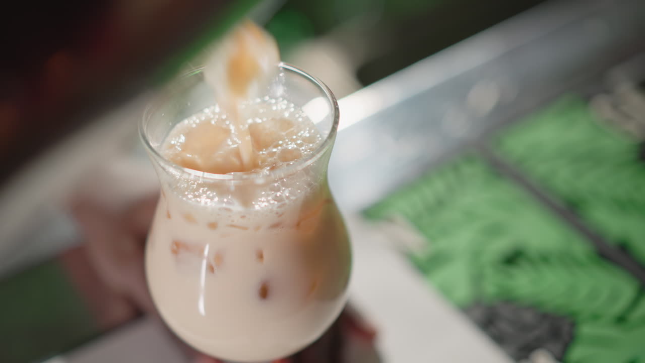Bartender pouring creamy iced drink from shaker into glass with ice cubes on bar counter. The drink is foaming, and background lights add a vibrant bar atmosphere