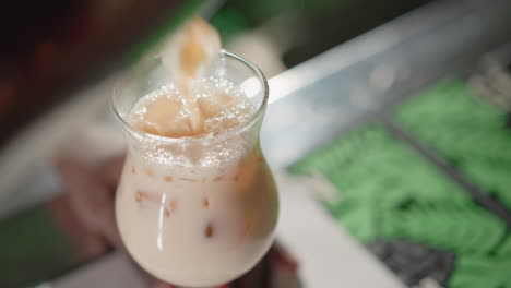 bartender pouring creamy iced drink from shaker into glass with ice cubes on bar counter. the drink is foaming, and background lights add a vibrant bar atmosphere