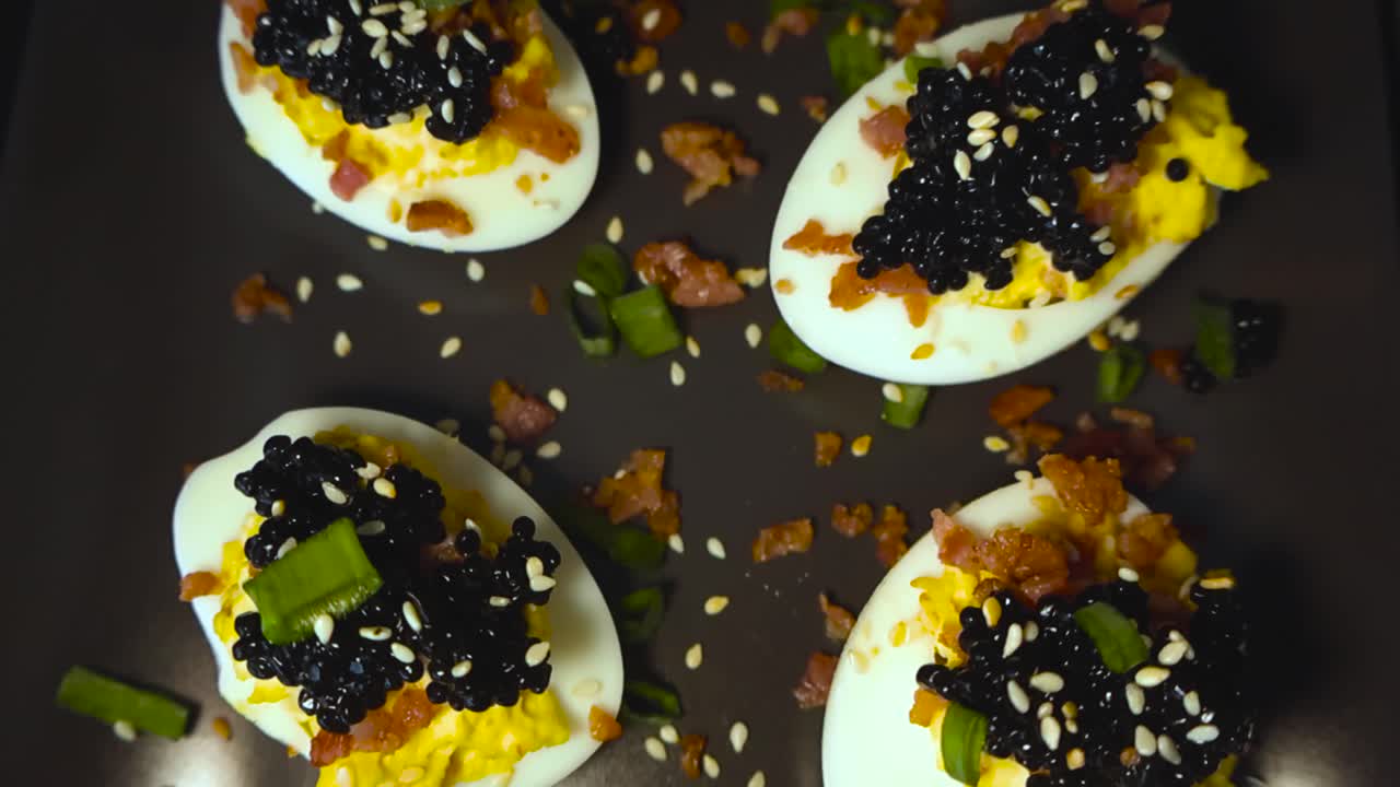 Ovenfra, filmisk glidende billede af gourmet fyldte æg toppet med sort kaviar, sprødt bacon, sesamfrø og friske krydderurter på en mørk tallerken.