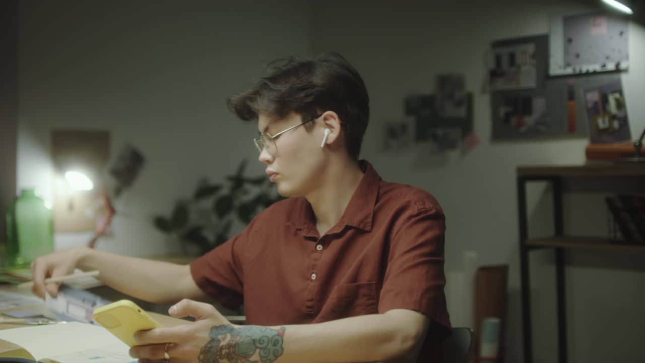 A young man studying or working at his desk in a dimly lit room, using a smartphone