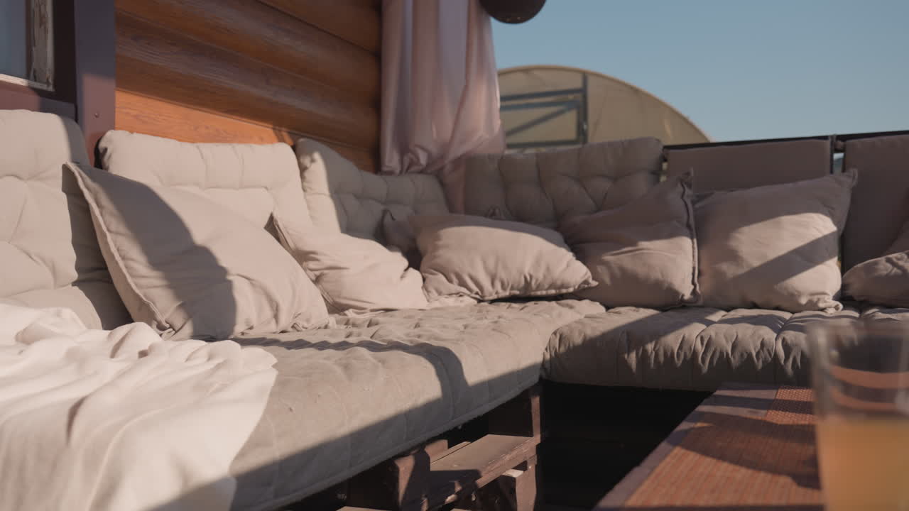 Sunlit Cabin Deck With Plush Sofa, Layered Cushions And Draped Blanket, Glass On Low Table, Warm Afternoon Light Casting Soft Shadows Across Log Wall And Curtained Window, Sky Visible Beyond Railing