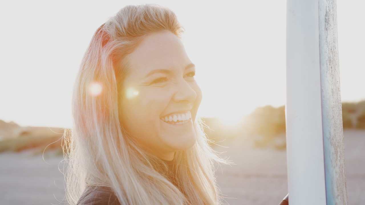 Woman Wearing Wetsuit Holding Surfboard Enjoying Surfing Vacation On Beach As Sun Sets