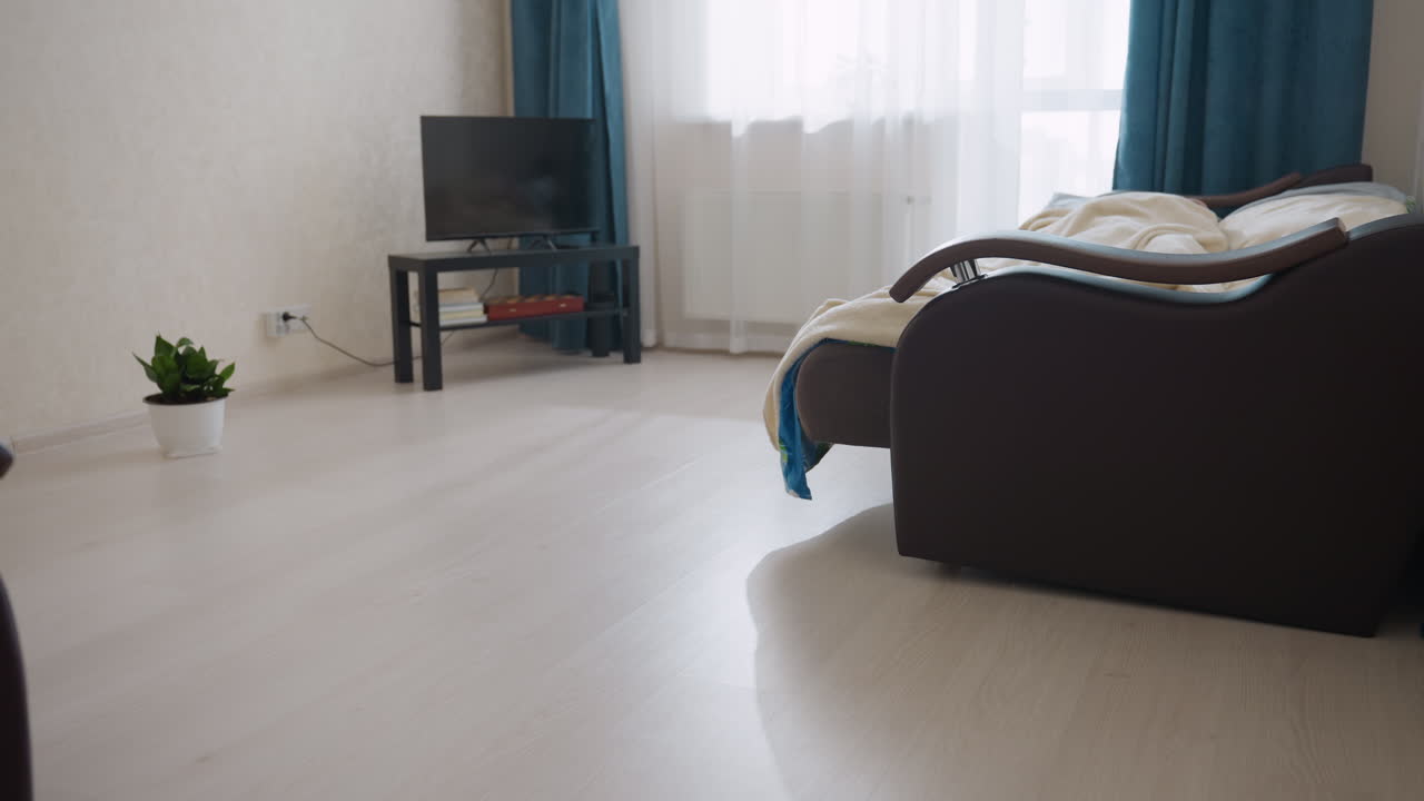 Individual sleeping under blanket rolling on bed in cozy bright bedroom with TV on black table near window and green potted plant on floor creating peaceful calm lifestyle