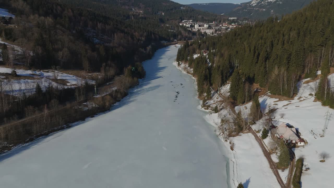 Drone flying in a snowy mountain valley over a frozen lake towards the Spindleruv Mlyn, the best ski area in Czechia.