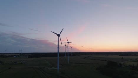 Aerial-establishing-view-wind-turbines-generating-renewable-energy-in-a-wind-farm,-evening-after-the-sunset-golden-hour,-countryside-landscape,-high-contrast-silhouettes,-drone-shot-moving-backward