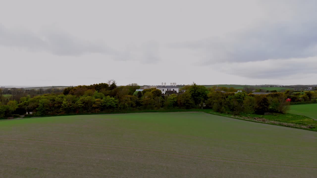 Luftfoto af en grøn mark i Belfast, der grænser op til tætte træer med fjerne bygninger og bølgende landskab under en overskyet himmel