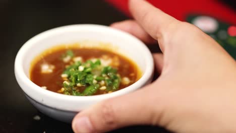 close-up of a hand holding a small bowl filled with sauce and green garnish.