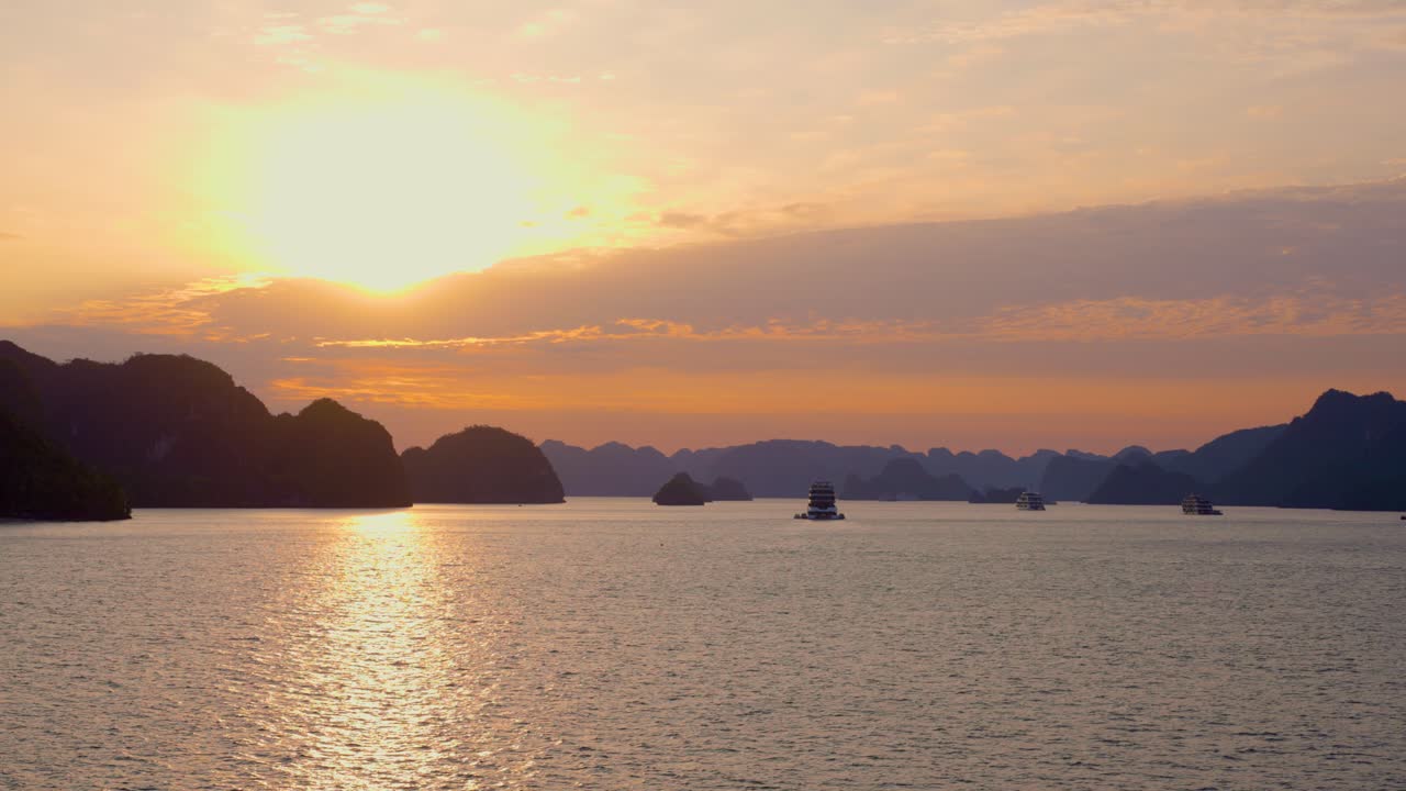 A mesmerizing sunrise bathes Ha Long Bay's limestone karsts in golden light, casting long shadows across tranquil waters where cruise ships drift serenely among the rocky islands