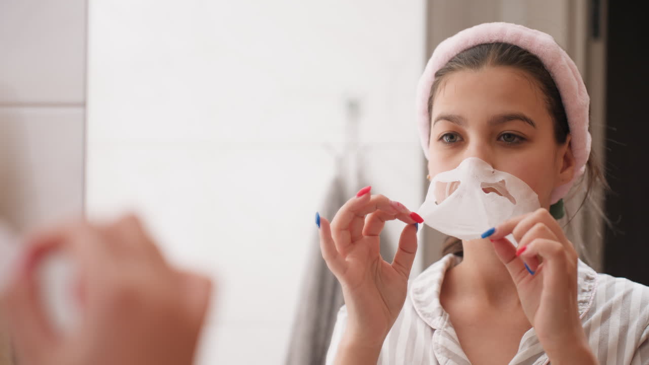 Girl Applies Mask With Smile, She Peels Lip Strip In Mirror, Young Woman Trying New Beauty Experiment Happily, Woman With Colorful Nails Enjoys Playful Skincare Moment In Front Of Mirror