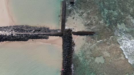 Top-view-aerial-of-rock-water-barrier-in-Waikiki-beach,-tilt-up-reveals-city