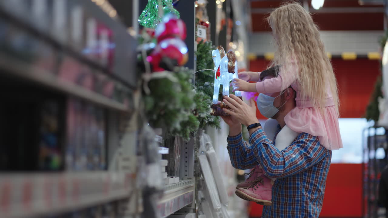 un padre camina con una máscara protectora por la tienda con su hija sobre sus hombros en un supermercado y elige un caballo brillante