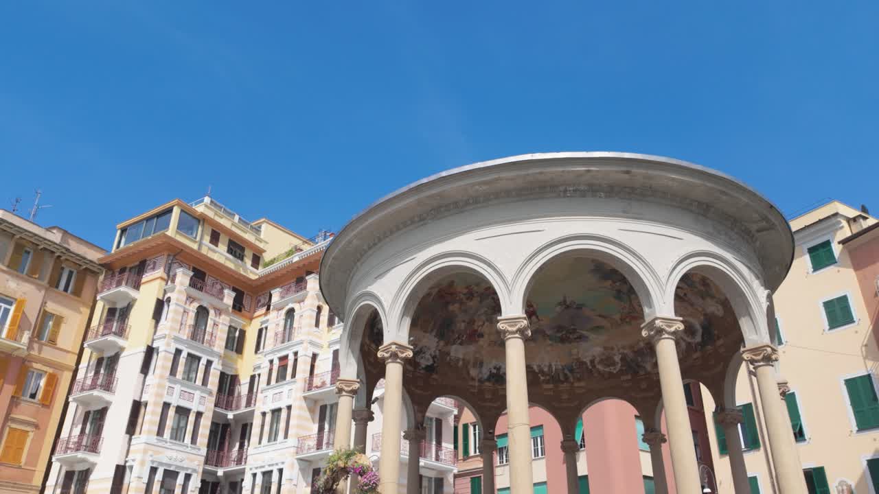 Riprese cinematografiche grandangolari del chiosco della musica ornato di Rapallo, sullo sfondo di colorati edifici storici sotto un cielo azzurro e senza nuvole. Facciate illuminate dal sole, tonalità vivaci e la struttura sottolineano il fascino italiano