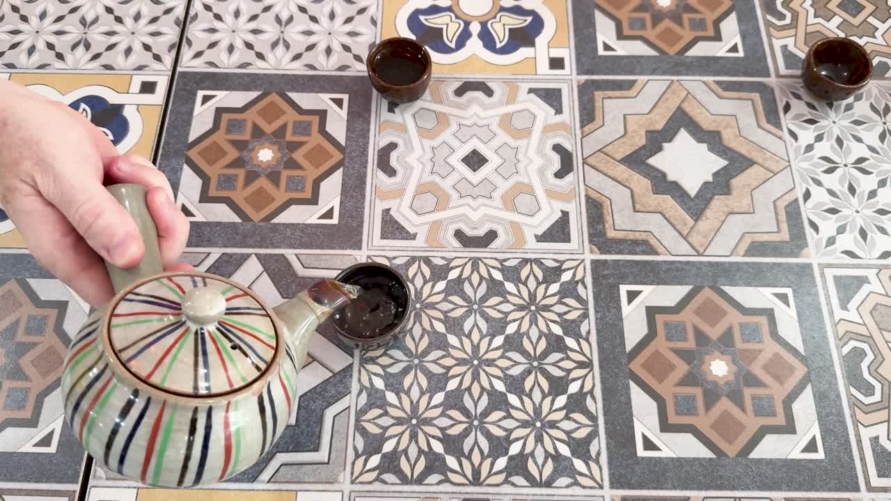Pouring tea from striped teapot into small cup on colorful patterned tile table, overhead view