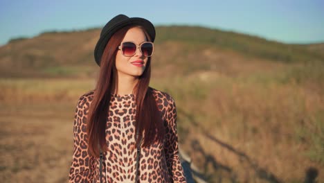 Joven-Mujer-Caucásica-Con-Sombrero-Negro-Y-Gafas-Caminando-Hacia-Y-Mirando-La-Cámara-En-El-Campo-Rural,-Retrato-De-Mano-De-Cerca