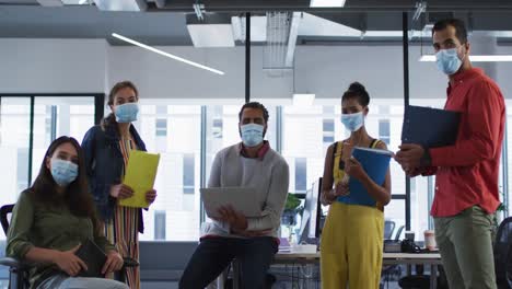 portrait of diverse group of work colleagues wearing face mask holding laptop, tablet and documents