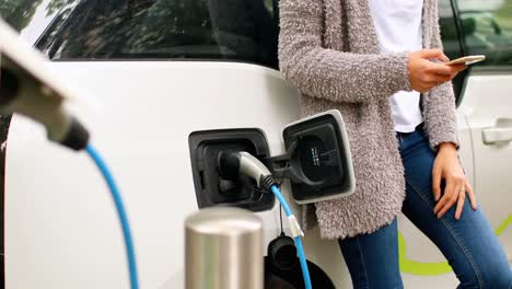 mujer usando teléfono móvil mientras carga un coche eléctrico en la estación de carga 4k