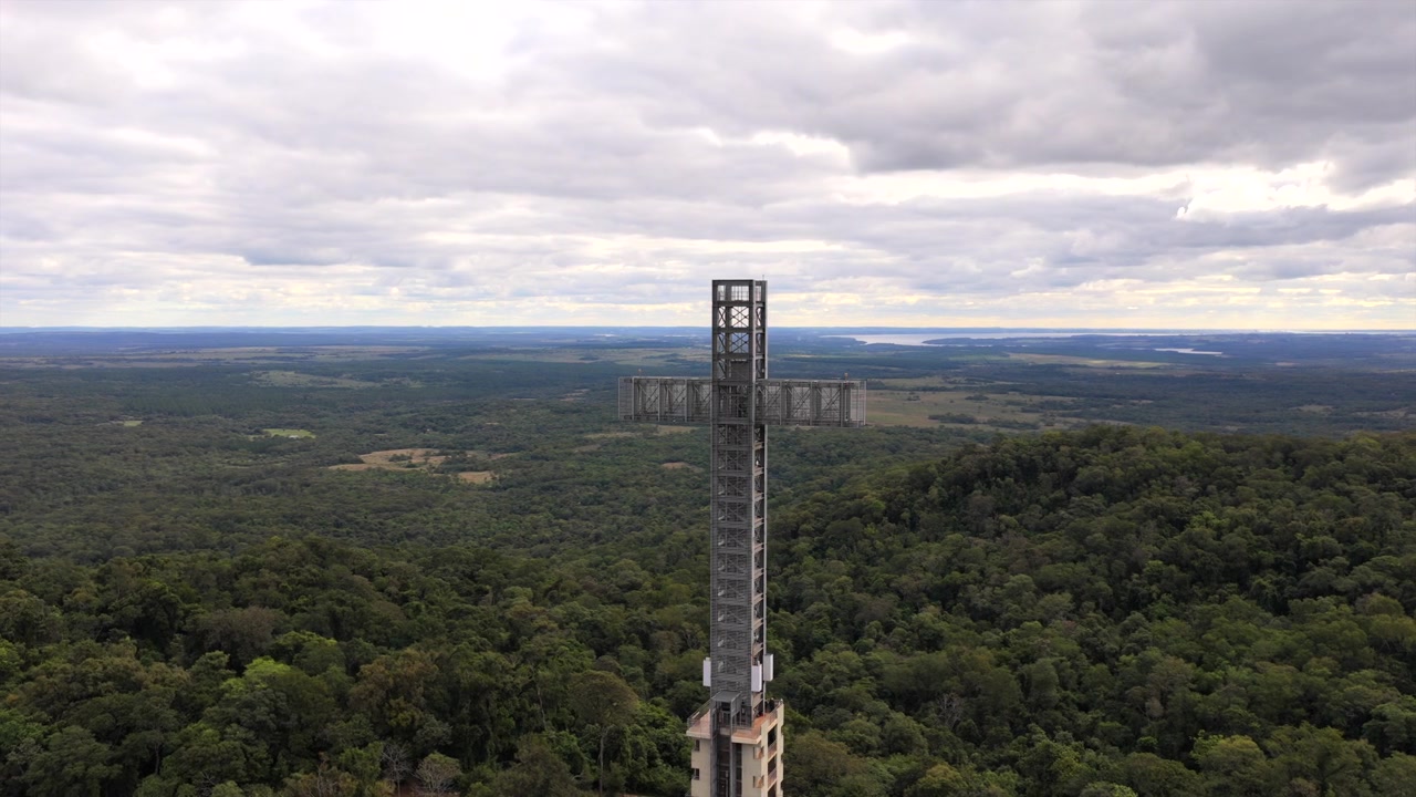Torre monumental em forma de cruz eleva-se sobre a paisagem da Mata Atlântica no Parque Temático de la Cruz, Santa Ana, Província de Misiones, Nordeste da Argentina, em uma tomada de estabelecimento em órbita de drone sob nuvens subtropicais.