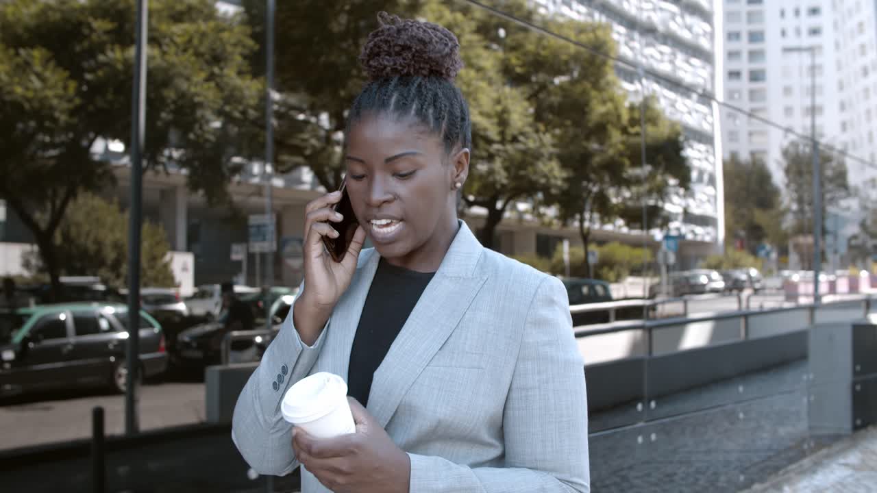 Dolly shot of a smiling African-American businesswoman walking outside, holding coffee and talking on mobile phone