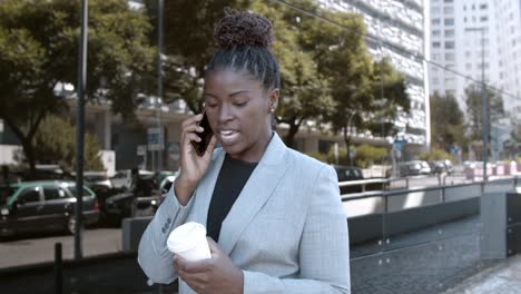 dolly shot of a smiling african-american businesswoman walking outside, holding coffee and talking on mobile phone