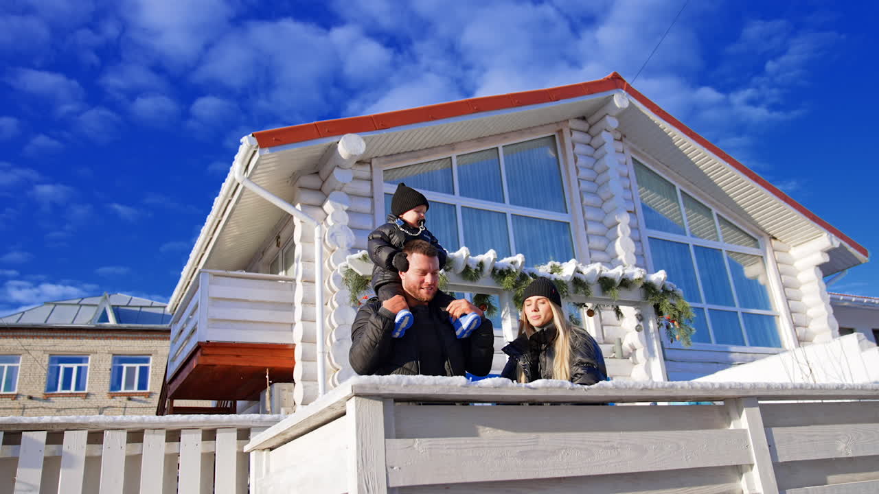 Loving family with little baby stand near their house on a sunny winter day. Toddler kid sucking a pacifier sits on dad's shoulders. Low angle view.