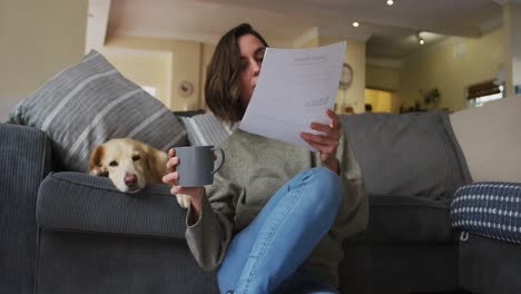 caucasian woman reading documents, working from home with her pet dog on sofa next to her