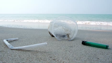 trash on a white sand beach, a lighter and plastic straw and cup lay close to the ocean