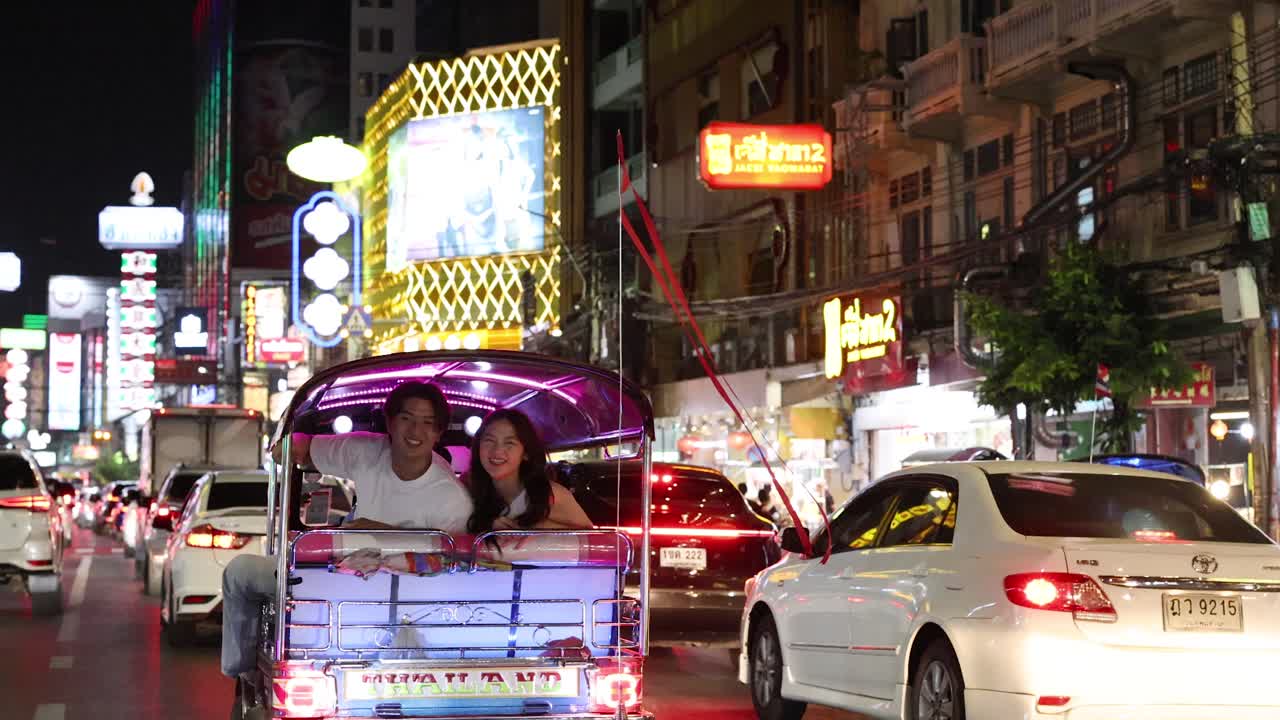 Smiling couple rides tuk tuk at night, colorful neon lights illuminate busy urban street scene