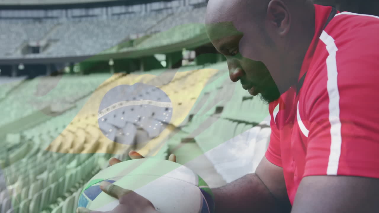 Uomo che tiene un pallone da calcio, con maglia rossa e sfondo di seggiolini dello stadio.