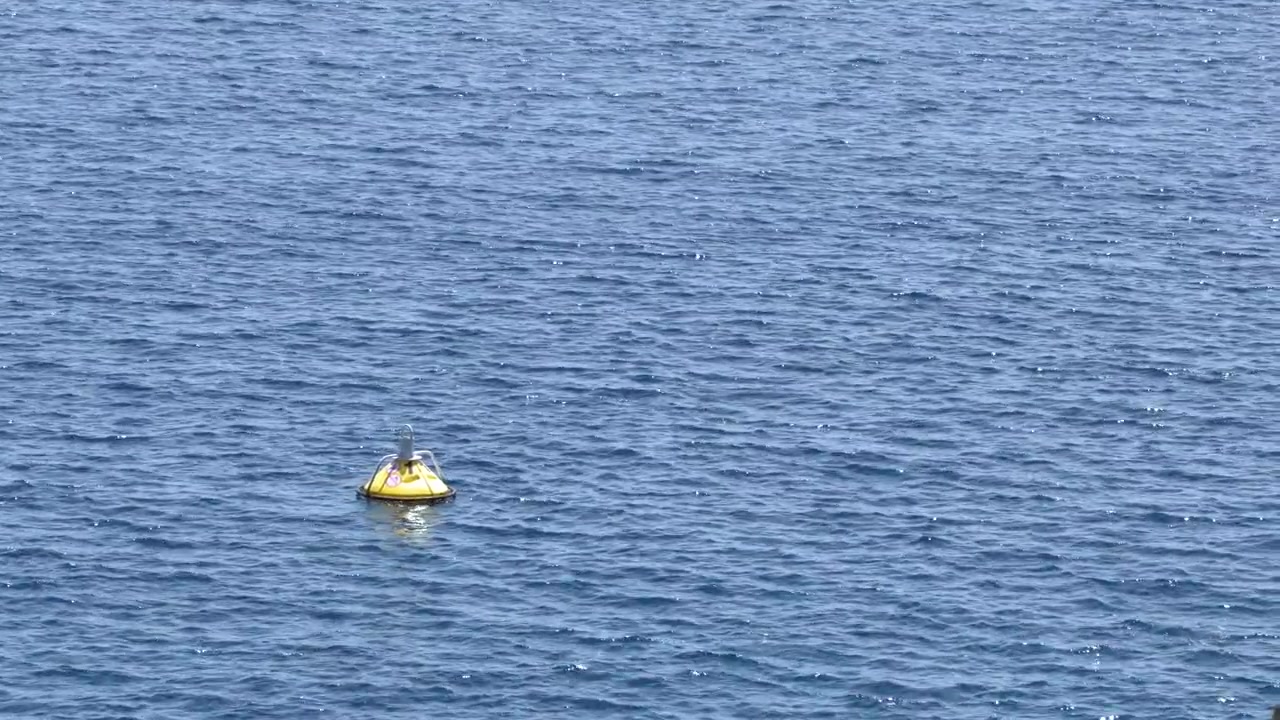 A yellow navigational buoy floats on sparkling blue sea water under bright natural sunlight.