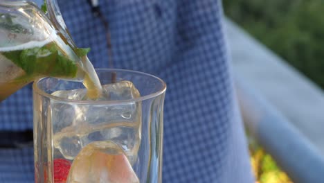 Slow-motion-pour-of-fruity-orange-alcoholic-drink-with-leaf-and-raspberry-from-clear-jug-into-ice-filled-glass-filling-up-on-summer-day