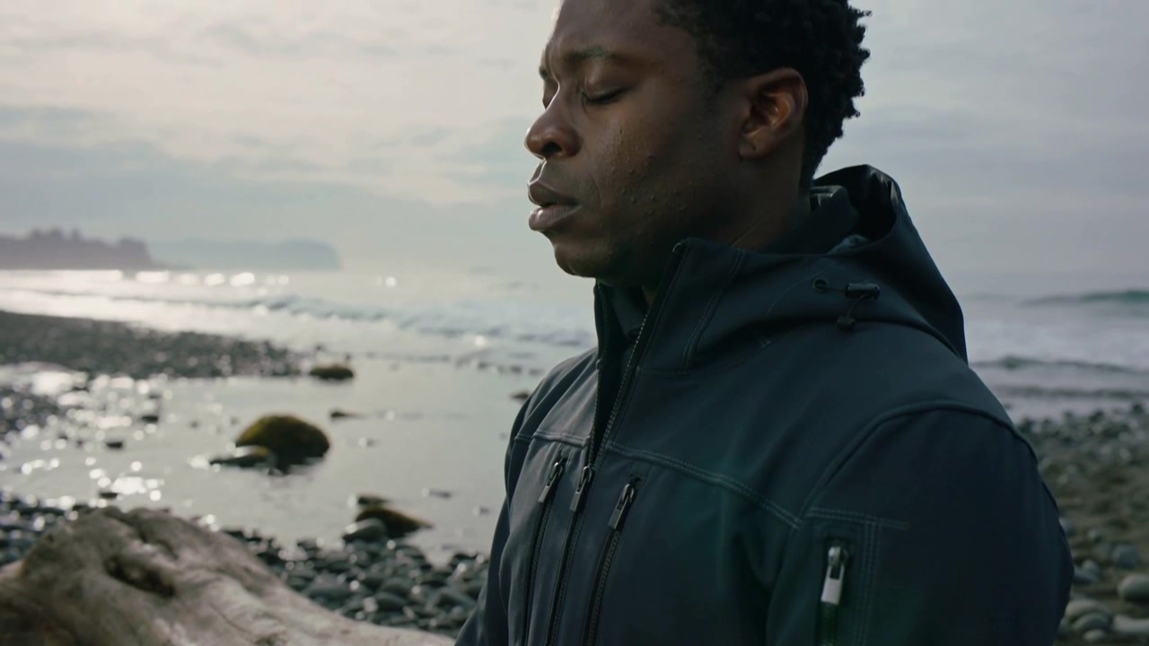 Sitting on driftwood log man turning head to sea as camera approaching, composing, hooded jacket
