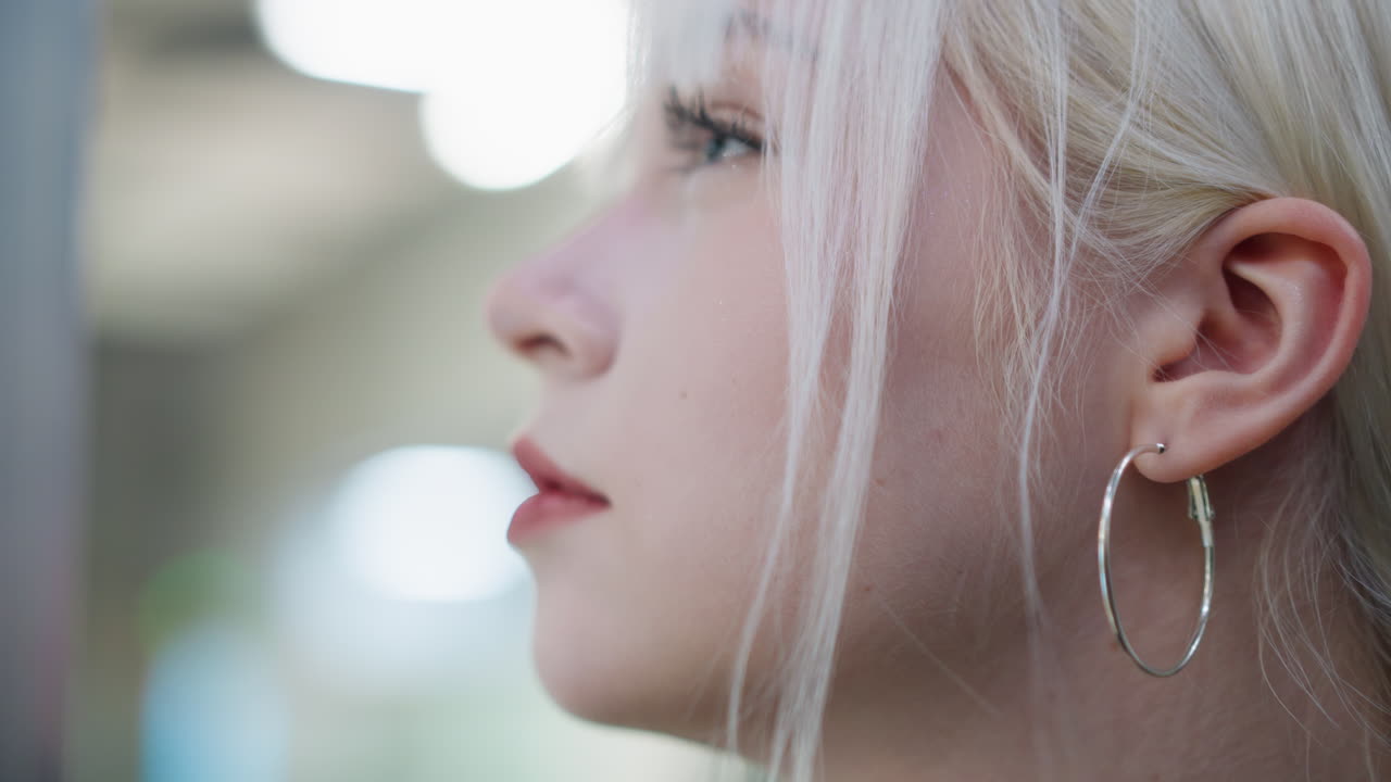 Close up profile of young blonde woman with hoop earring looking focused and thoughtful, side view highlighting natural features, soft indoor lighting creating calm and reflective mood