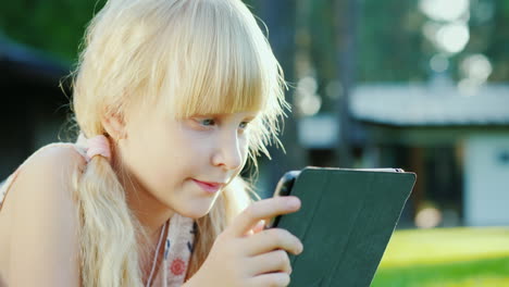 Porträt-Eines-6-jährigen-Mädchens,-Das-Auf-Einem-Tablet-Spielt,-Liegt-Auf-Dem-Gras-In-Der-Nähe-Des-Hauses-4k-10-Bit-Video