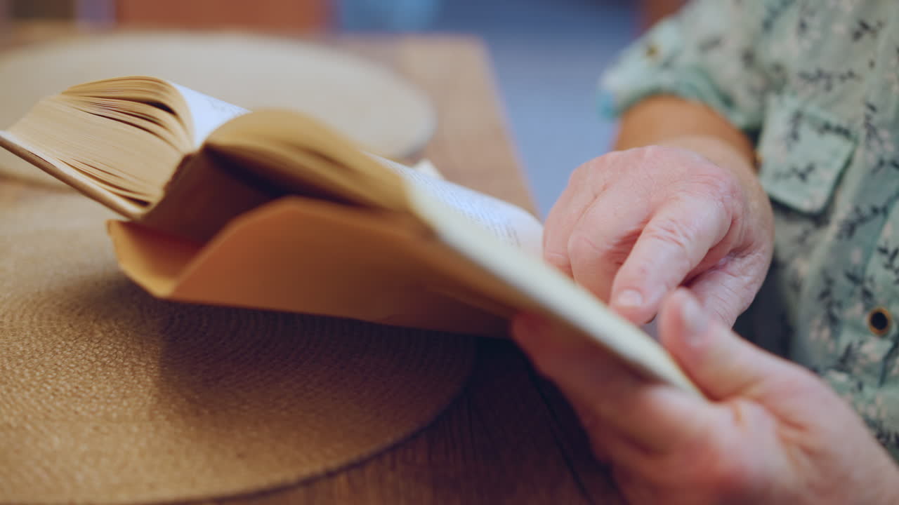 Mãos iluminadas pelo sol segurando livro aberto, avó sentada à mesa com chapéu de palha e camisa estampada visíveis, dedos virando páginas, brilho quente da tarde, clima suave de contação de histórias sugerindo leitura em voz alta
