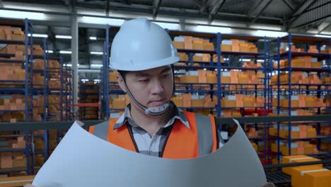 close up de ingeniero masculino asiático con casco de seguridad mirando el plano en sus manos y mirando a su alrededor mientras estaba de pie en el almacén con estantes llenos de bienes de entrega