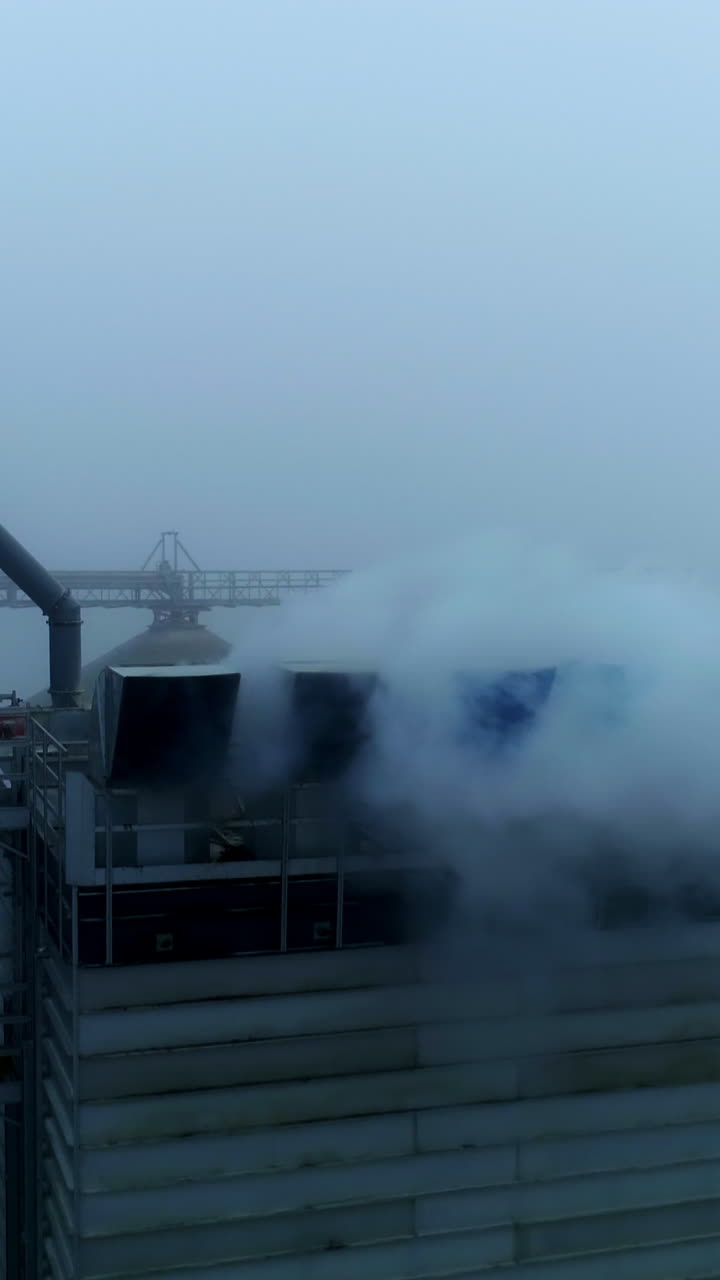 High metal constructions, supports and pipes at the modern elevator plant. Thick white smoke coming into camera. Grey foggy day backdrop. Vertical video