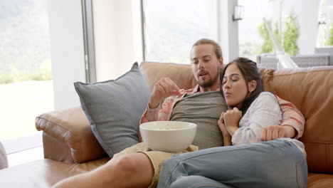 A-young-biracial-couple-relaxes-on-a-leather-sofa,-copy-space,-watching-a-movie-with-popcorn