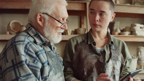 senior man and young woman using tablet and talking in pottery studio