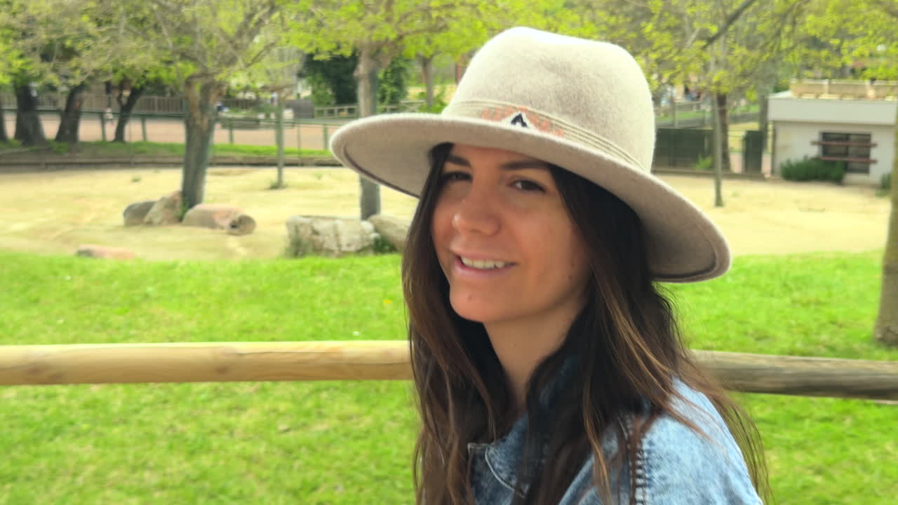 Close-up of a smiling young woman wearing a wide-brimmed hat and denim jacket, walking outdoors through a green park on a sunny day, capturing happiness and natural lifestyle