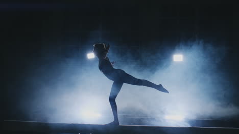 the girl is a professional athlete performs gymnastic acrobatic trick on a beam in backlight and slow motion in sports gymnastic clothing. smoke and blue. jump and spin on the balance beam