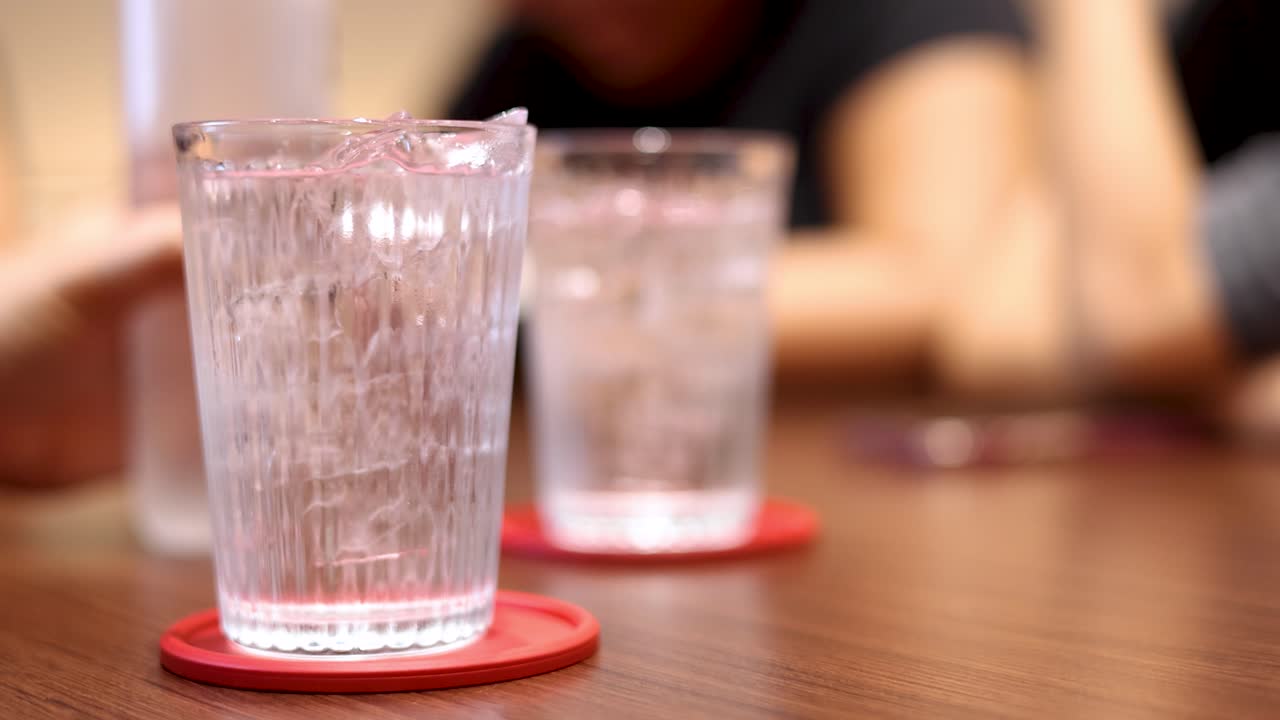 Hand pours water into ice-filled glass on wooden table in warm, social restaurant setting