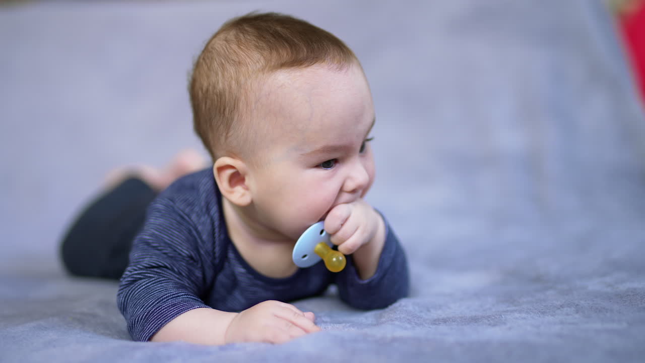 Tiny cute boy lies on bed and chews his pacifier. Little kid drops the nipple and starts to chew his fingers. Close up.