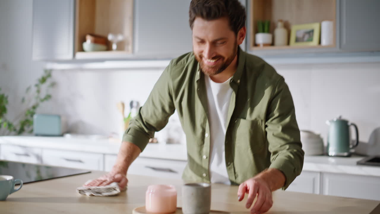 Uomo sorridente che pulisce il bancone della cucina con un panno. Ragazzo barbuto felice che pulisce il tavolo