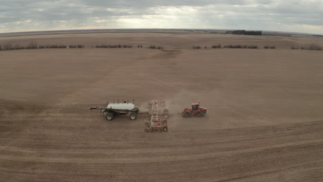 Vista-Aérea-Lateral-Panorámica-Sobre-La-Máquina-Sembradora-Industrial-Roja-Que-Tira-De-Fertilizante-En-Un-Campo-Agrícola-Plano-En-El-Campo-Rural-En-Un-Día-Nublado,-Corriente-Rápida,-Saskatchewan,-Canadá