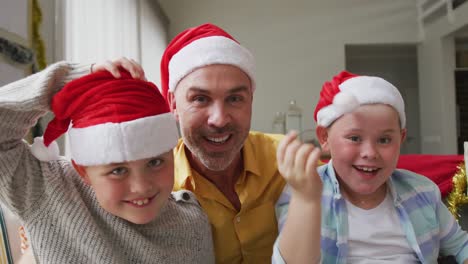Portrait-of-caucasian-father-and-two-sons-wearing-santa-hats-waving-and-smiling-sitting-on-the-couch
