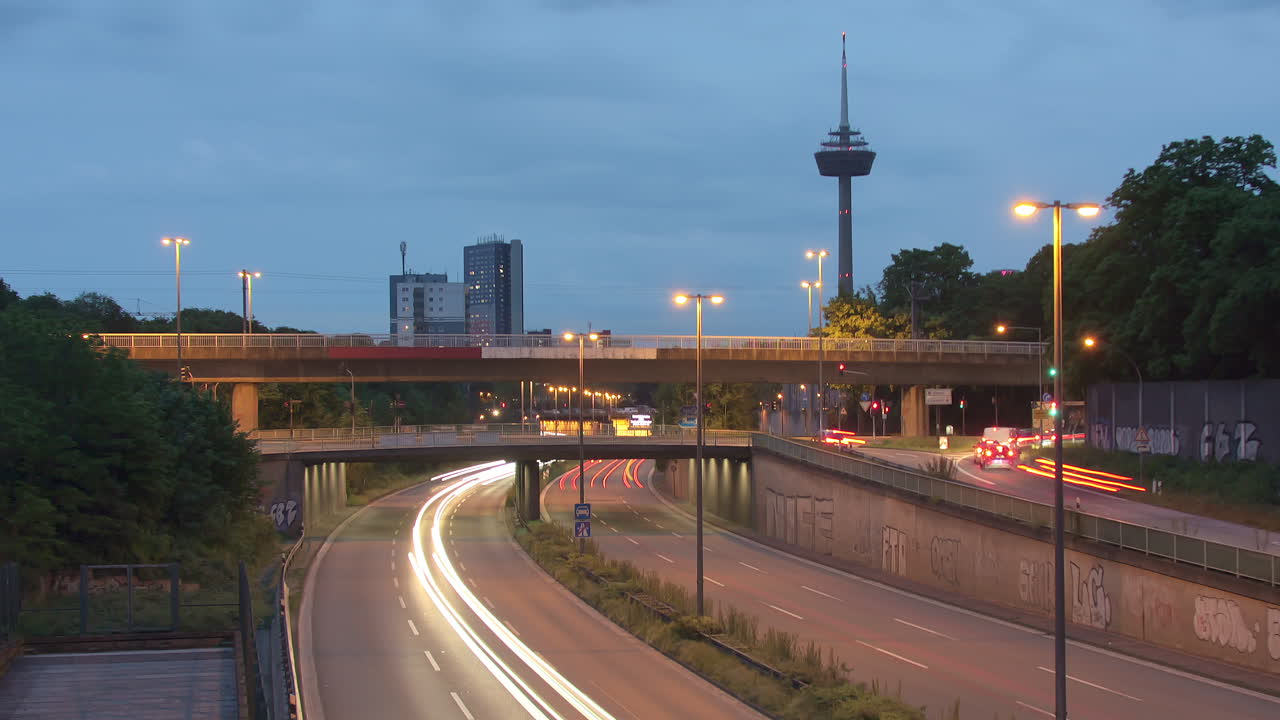 Traffic Flow and Cologne Skyline. Witness bustling cars with light strokes on a highway, a prominent TV tower, moving clouds, and Cologne's panoramic skyline in the evening. Energy and dynamism. Z003