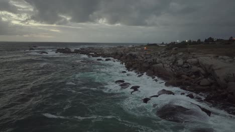 drone volando hacia atrás sobre el océano en camps bay beach en ciudad del cabo, sudáfrica. es un día nublado y las olas golpean la costa.